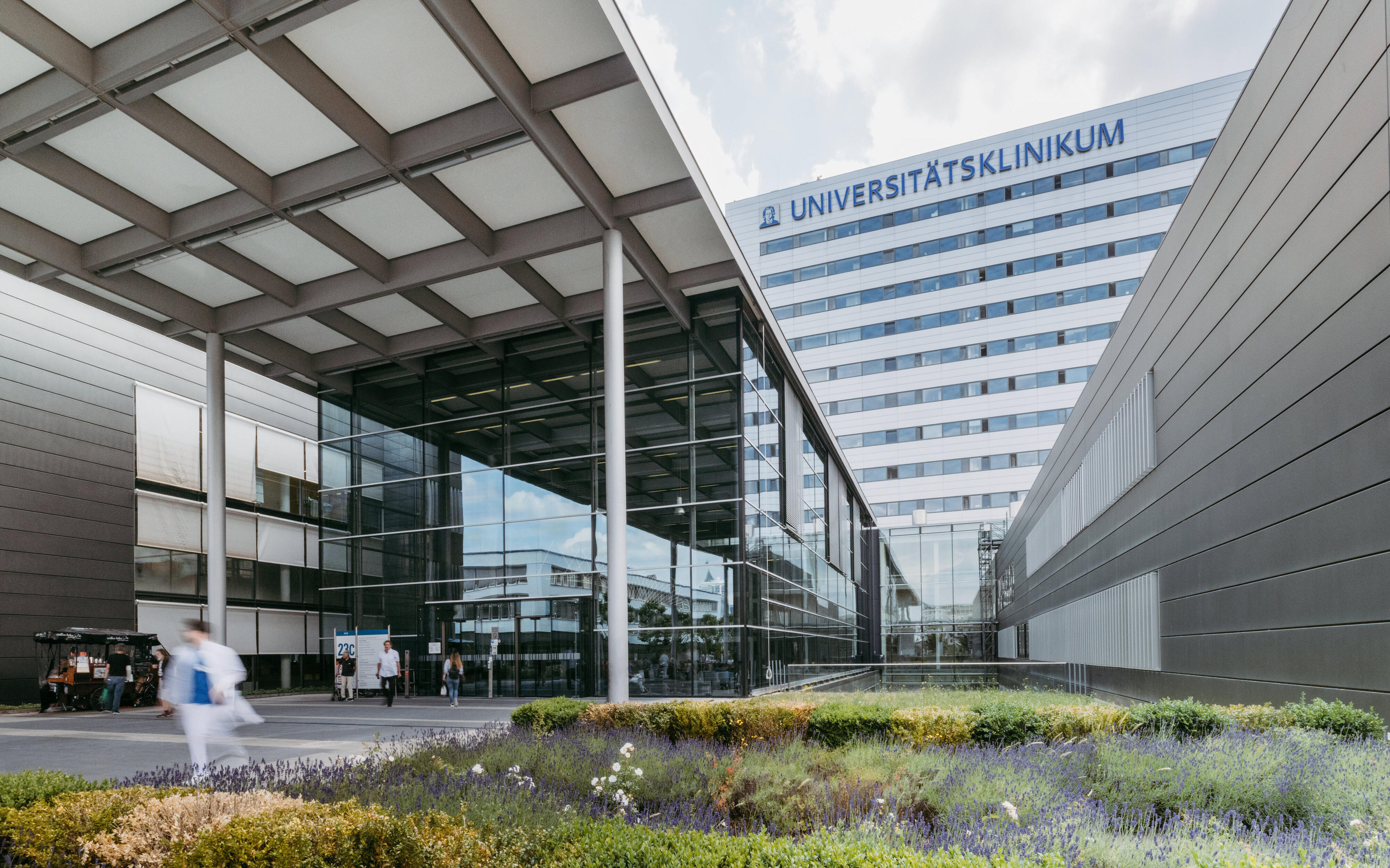 Eingangsbereich des Universitätsklinikums Frankfurt von außen, daneben ist Bepflanzung zu sehen. Teile der Fassade sind zudem sichtbar, wie der Schriftzug Universitätsklinikum